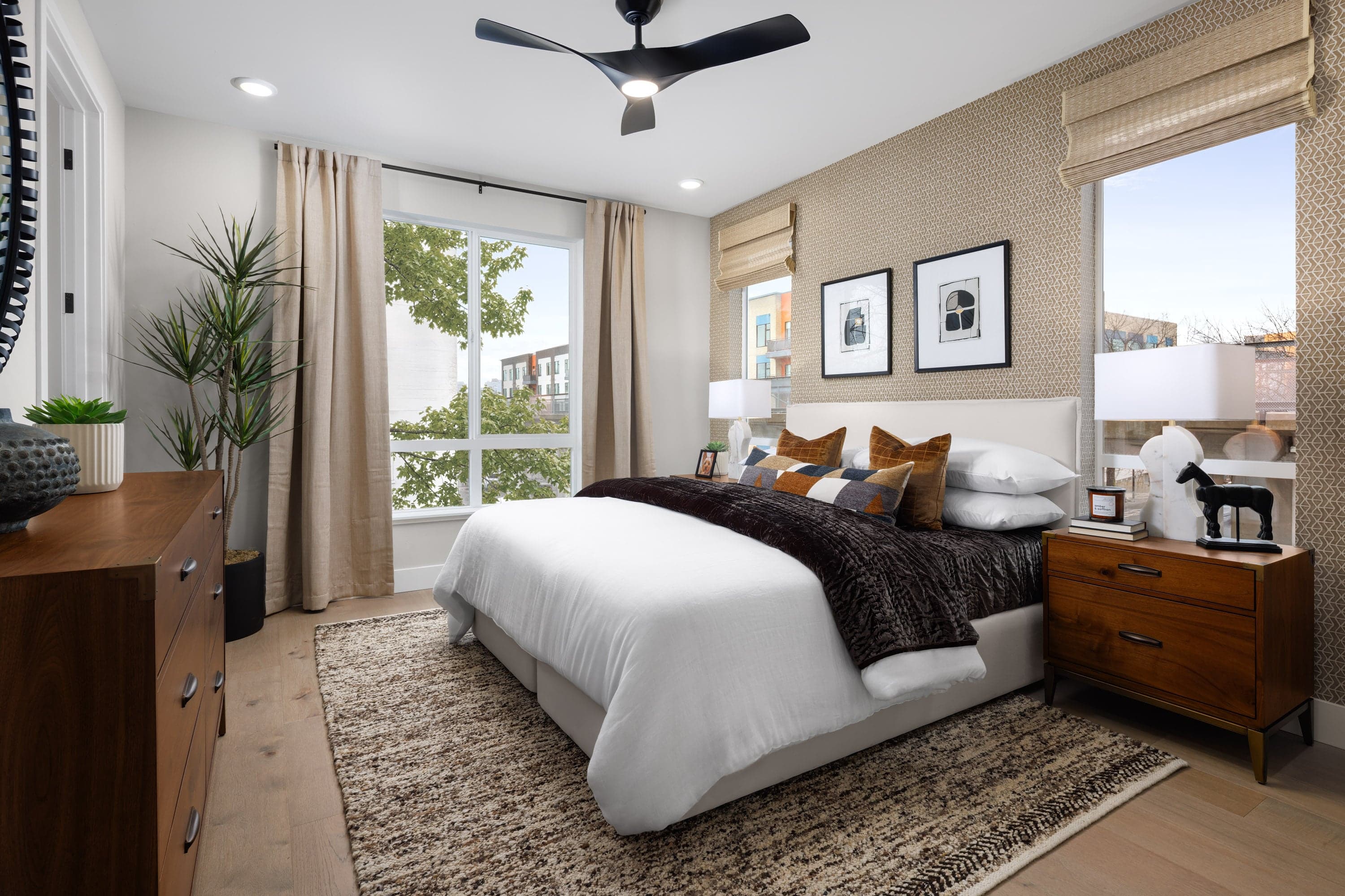 bedroom with tan patterned wallpaper behind white upholstered bed, wood floors, three windows, and black ceiling fan